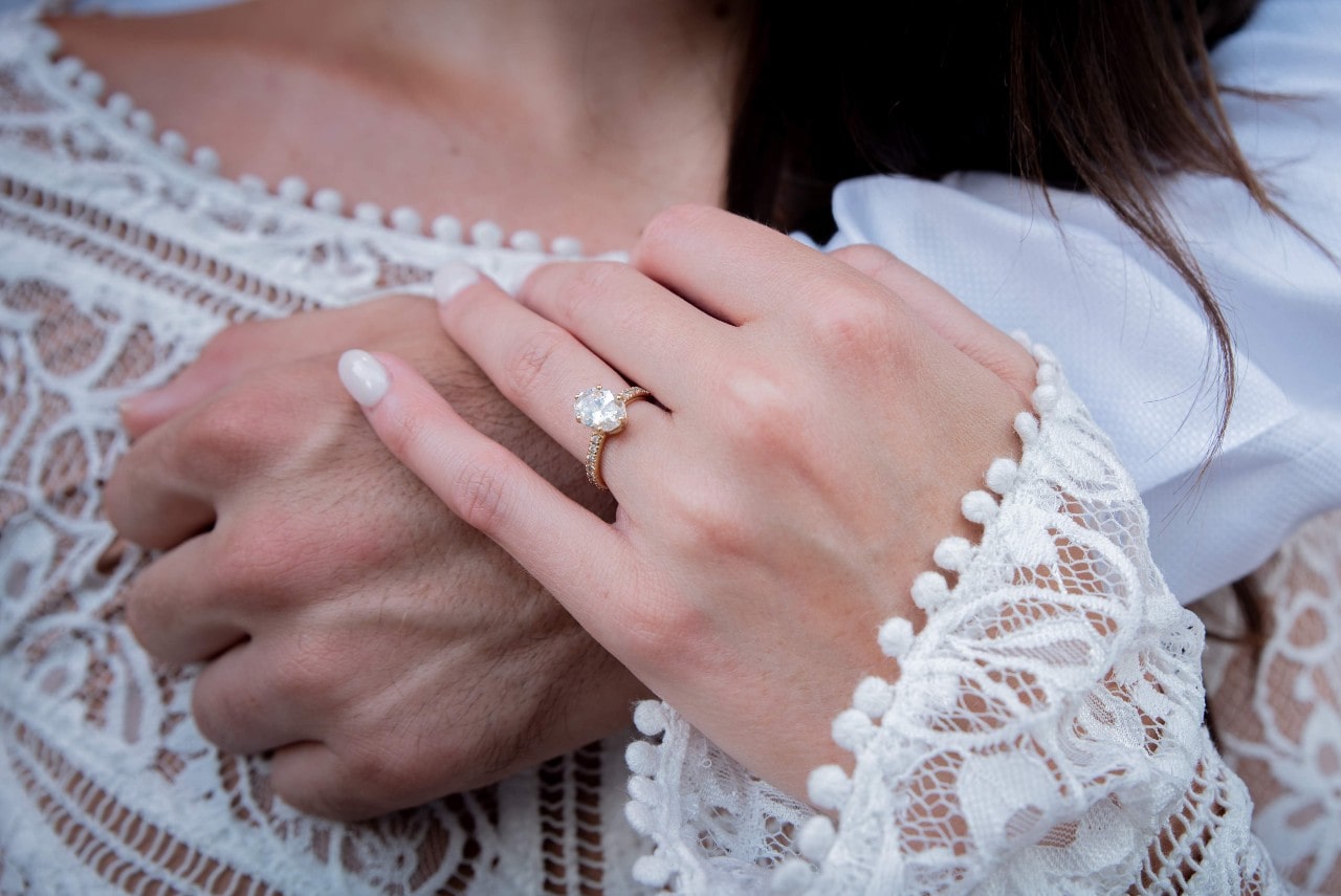 A bride holding the wrist of her love who has their arm around them, focus is on the oval center stone and side stone engagement ring