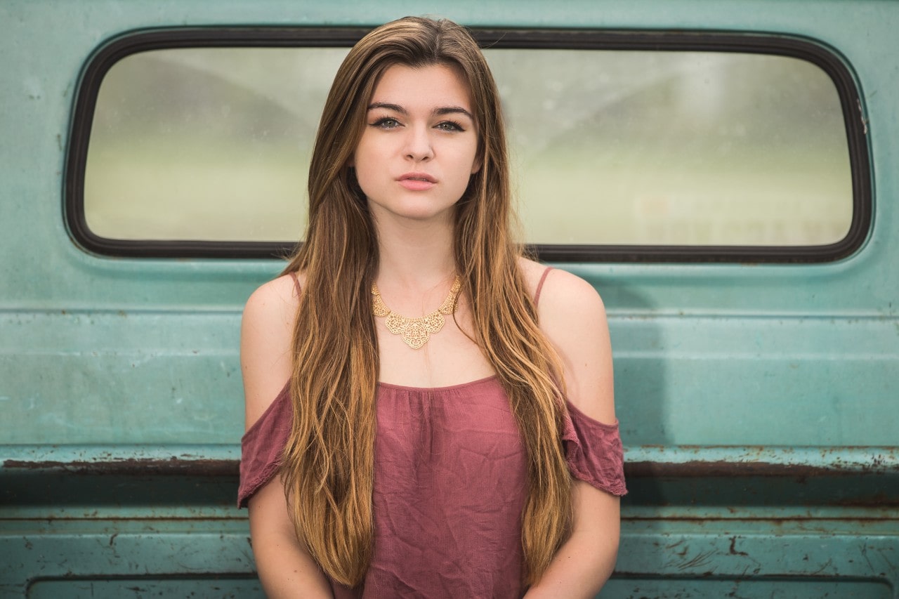 A woman with long hair and an elaborate necklace sits in the back of an old blue pickup truck.