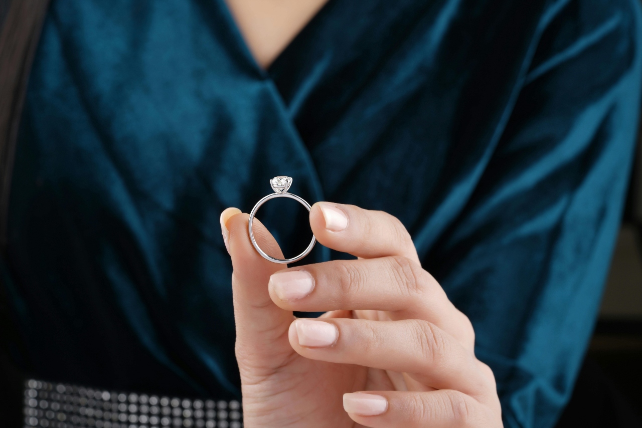 A woman in a blue velvet outfit holding up a solitaire engagement ring with a round diamond