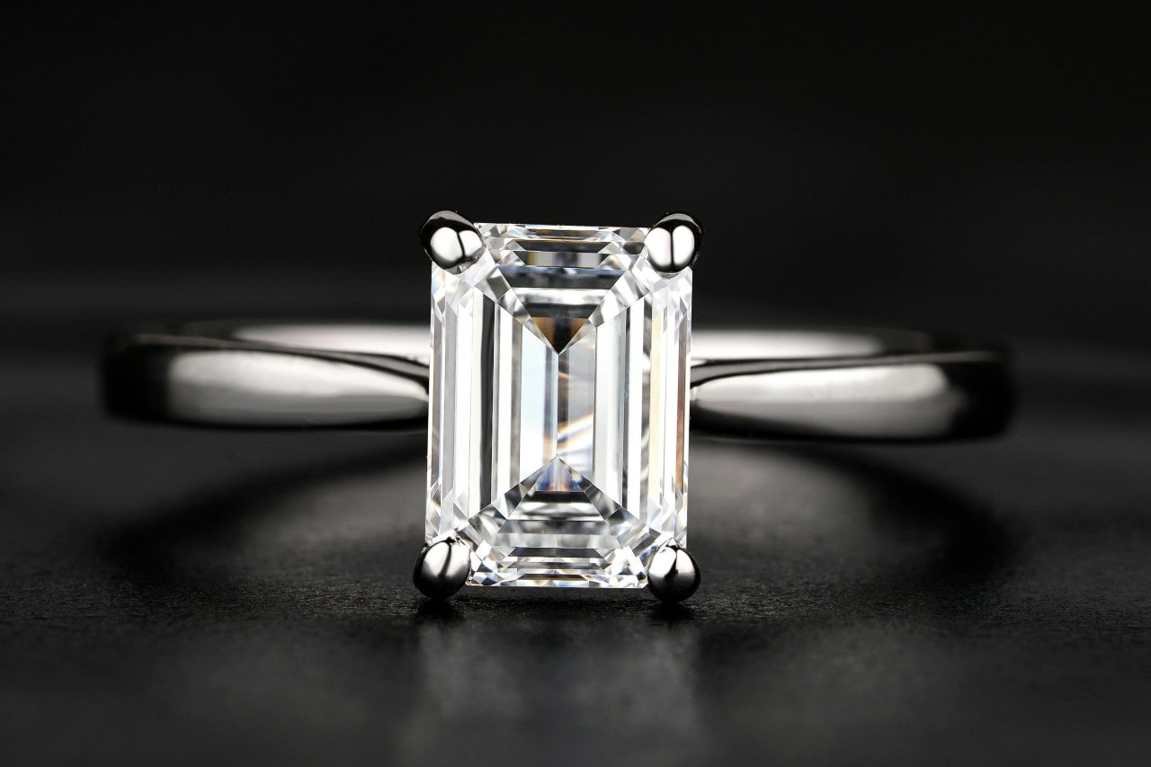 A close up of solitaire emerald cut diamond engagement ring on a black background