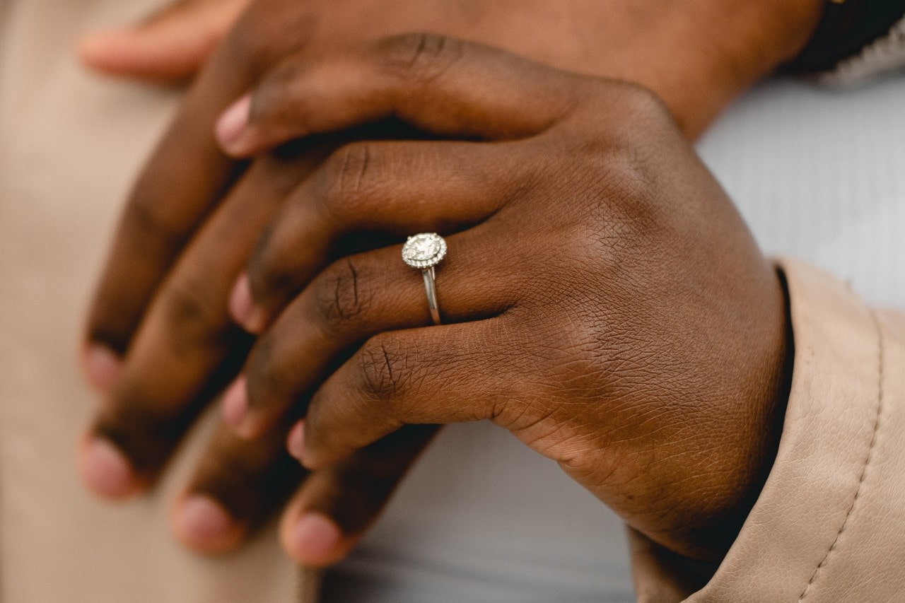 A couple holding hands, the woman wearing a silver, halo, oval cut engagement ring