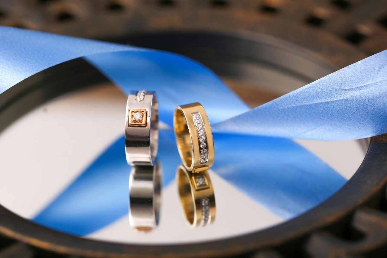 A close-up of two mixed metal wedding bands by a blue ribbon sitting on a mirror.