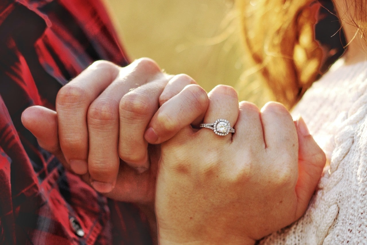 Close-up of couple hands gently clasp, with an emphasis on halo engagement ring against a softly blurred background.