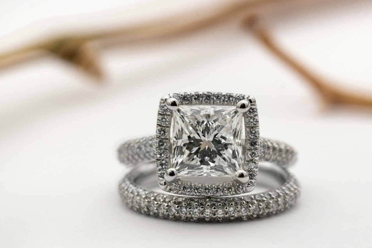 Princess-cut diamond engagement ring with halo and coordinating band on a blurred romantic background.