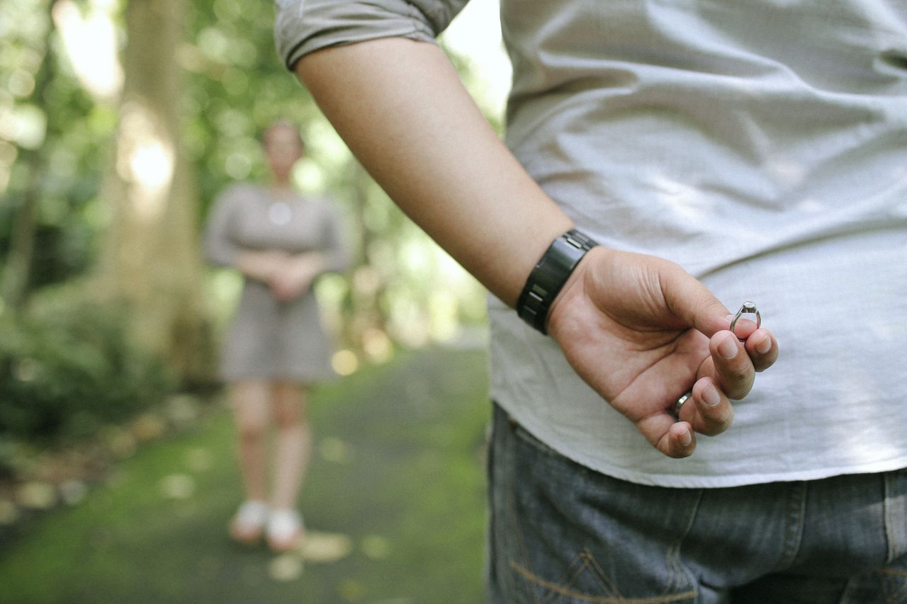 A man holding a round-cut engagement ring behind his back moments before proposing to his girlfriend