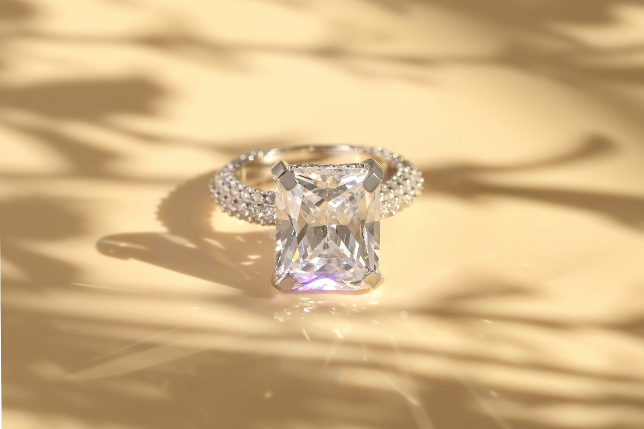 A radiant engagement ring with a cushion cut center stone and pavé side stones glistening against a soft, shadowed beige background.