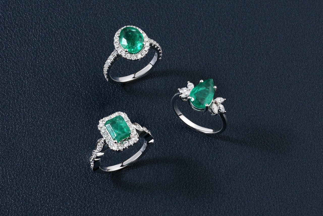 Three emerald engagement rings with sparkling diamond accents on a dark blue textured background.