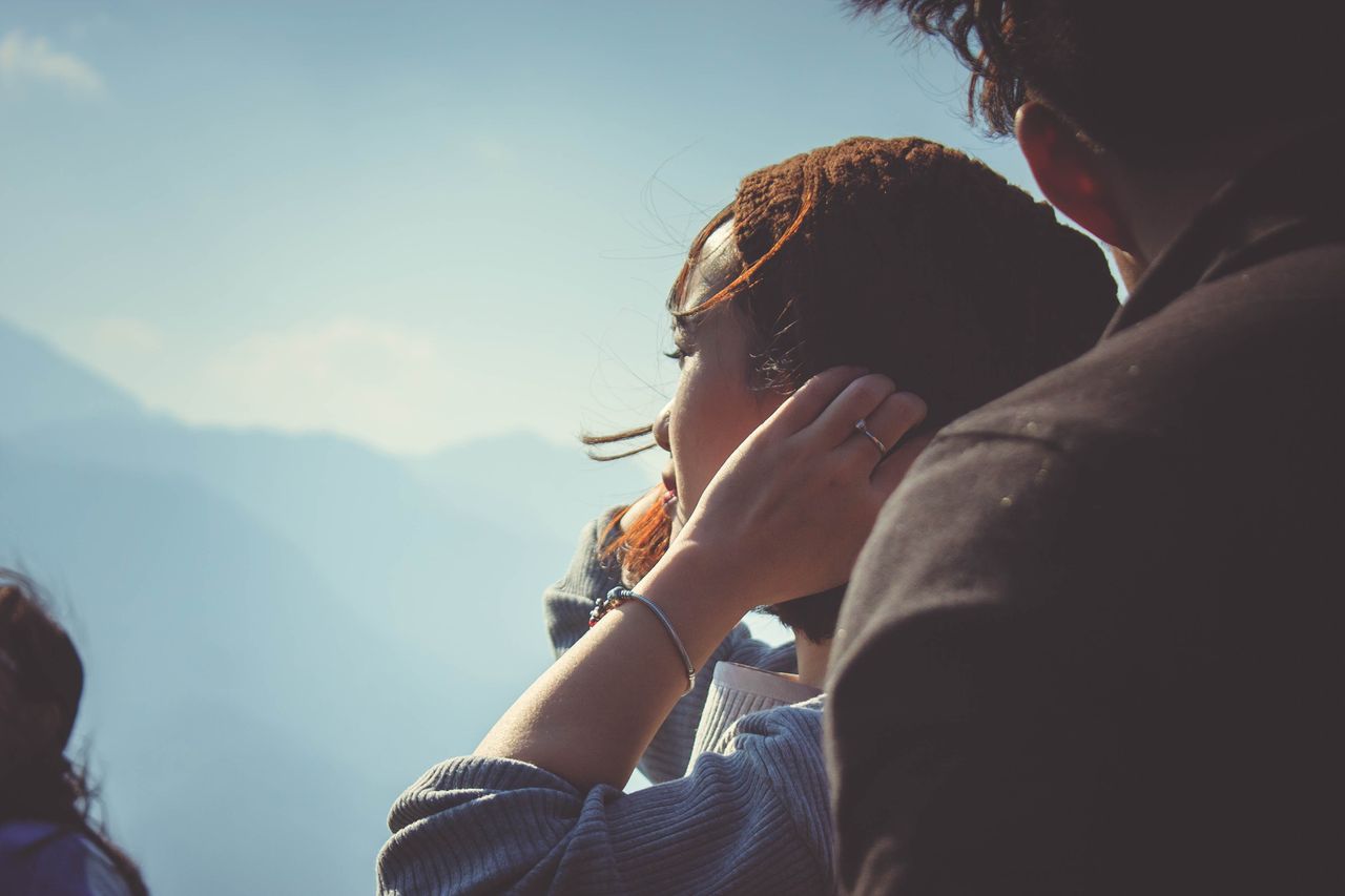 A man and woman up on a mountain enjoying the view, the woman holding her beanie and showcasing her silver bracelet and ring.