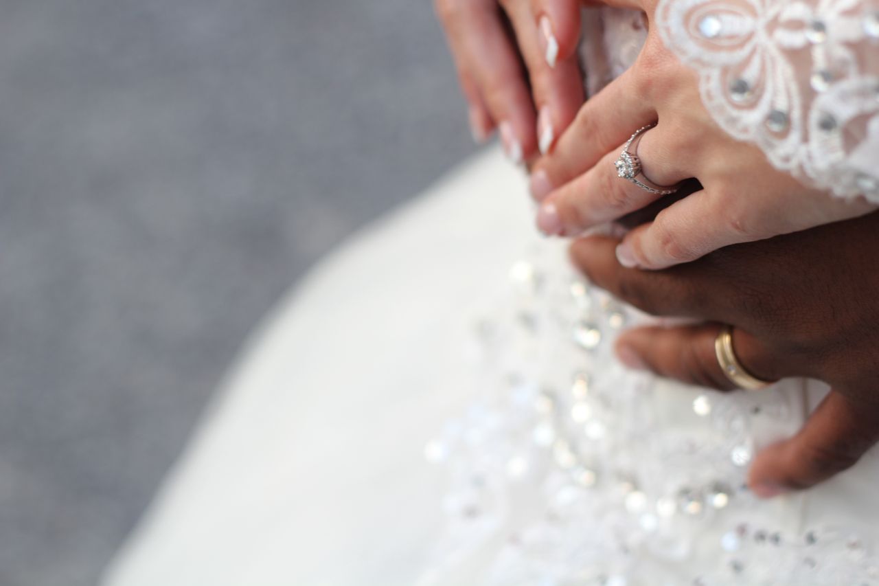 A bride with a halo ring rests her hand on top of her groom’s hands