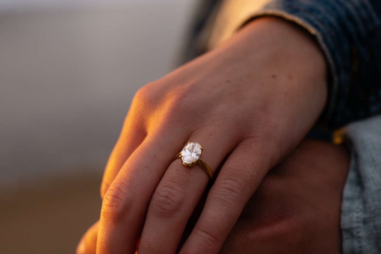 Image of a couple’s hands in fading sunlight, the woman wearing a gold, oval cut engagement ring