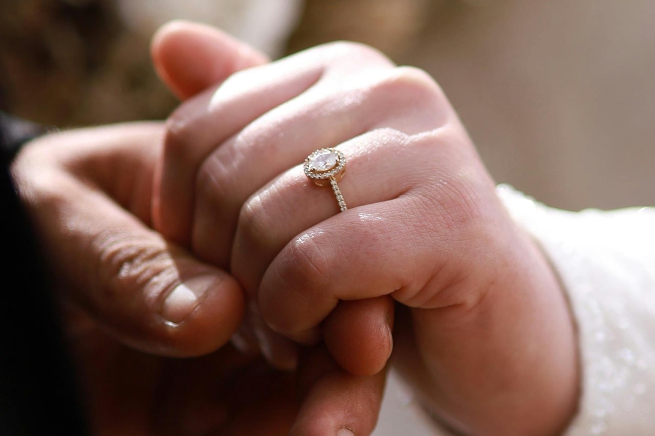 A close-up of two hands clasped together, with a bride wearing a halo engagement ring featuring a round diamond.