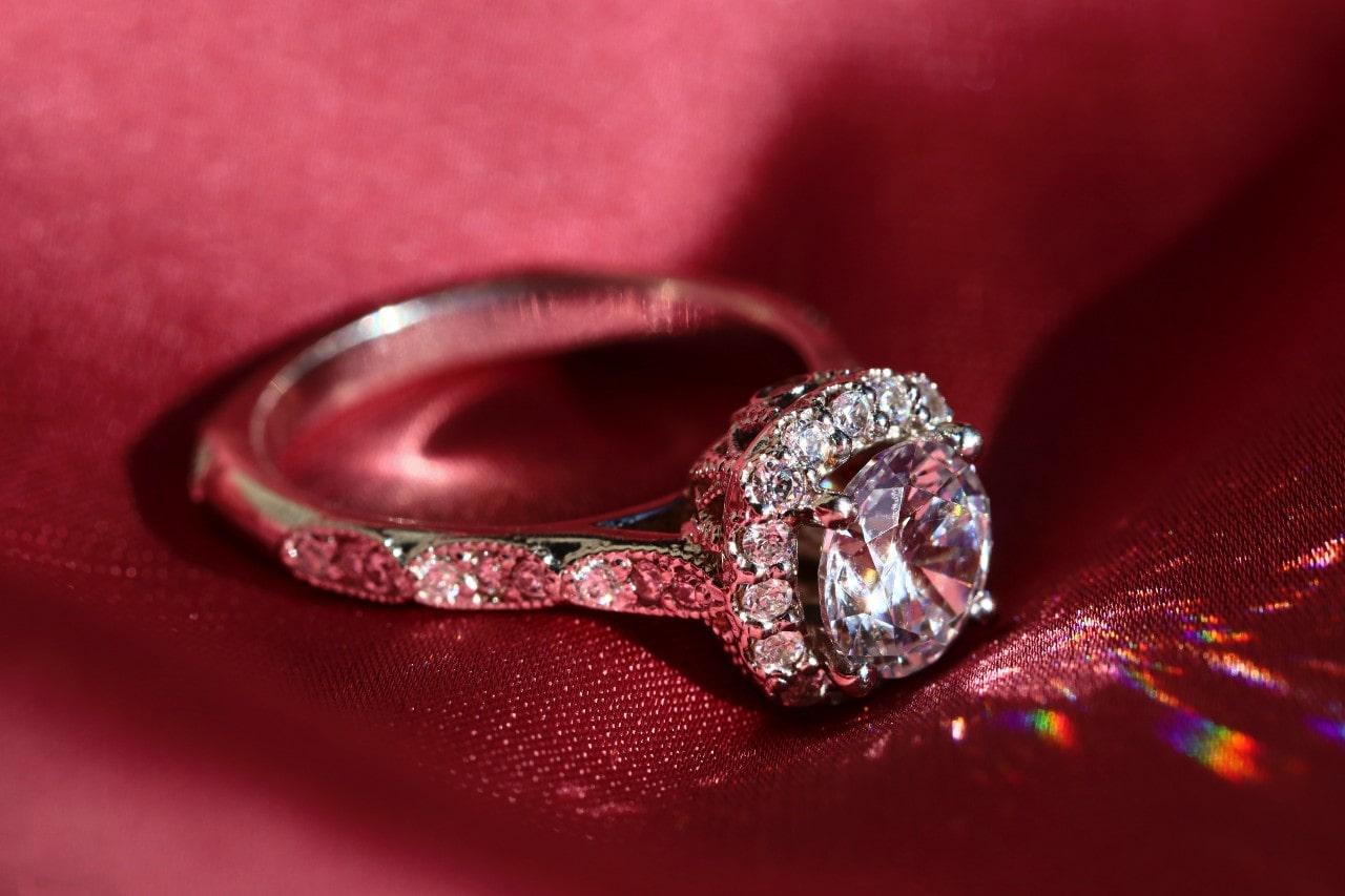 A sparkling diamond ring with a large central stone and diamond-encrusted band rests on luxurious red fabric.