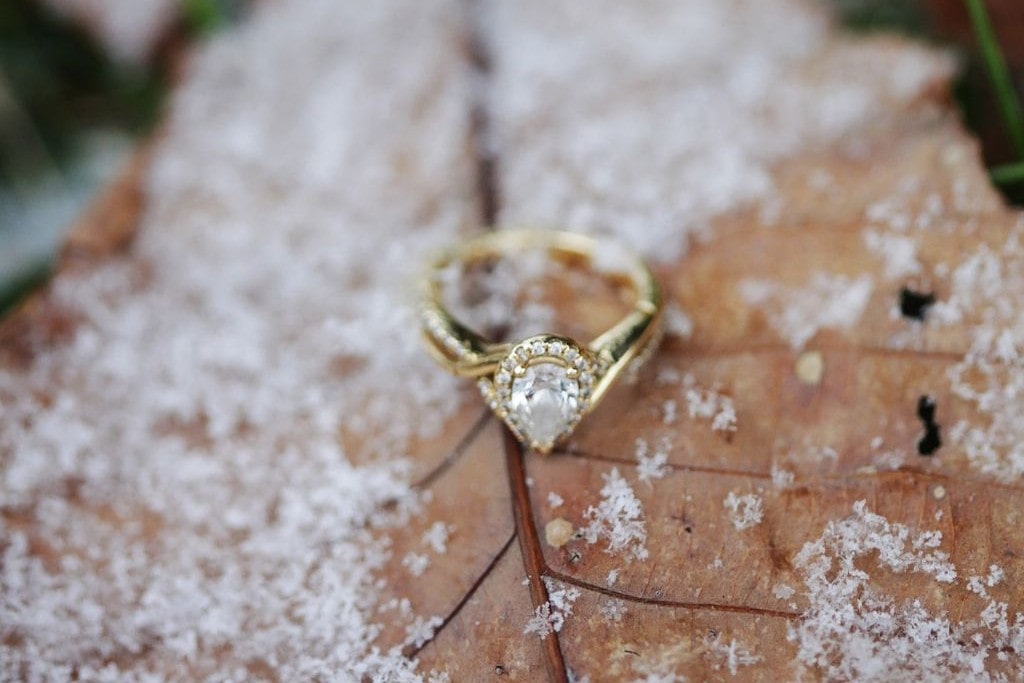 A pear-shaped halo engagement ring with a gold band rests on a brown leaf, dusted with snowflakes.