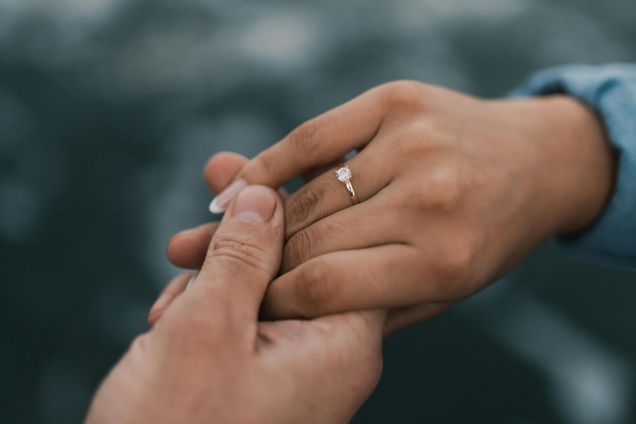 A close-up of a man holding his partner’s hand, a simple solitaire engagement ring on her finger.