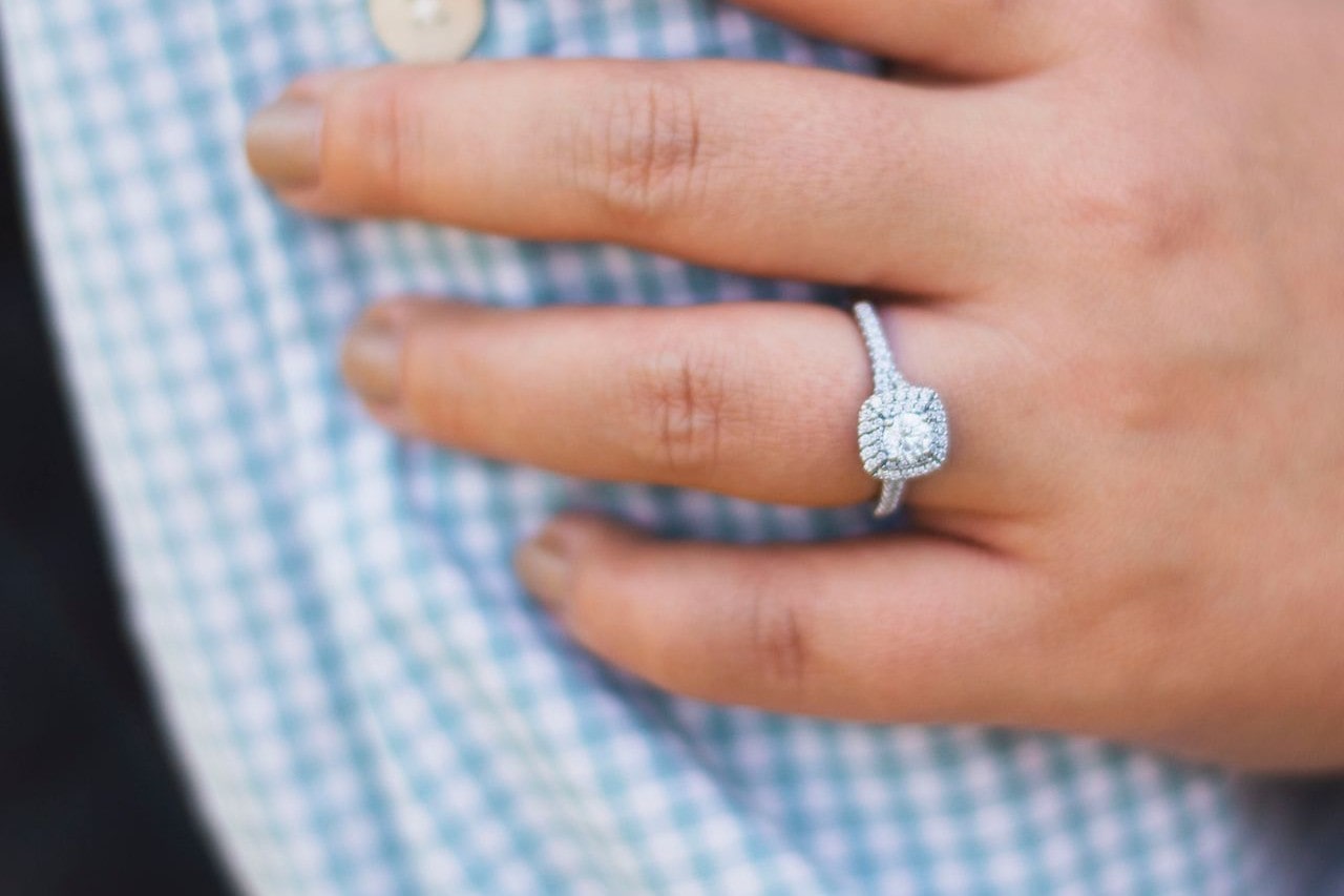 A hand with manicured nails rests on a blue plaid fabric, showcasing a sparkling diamond ring.