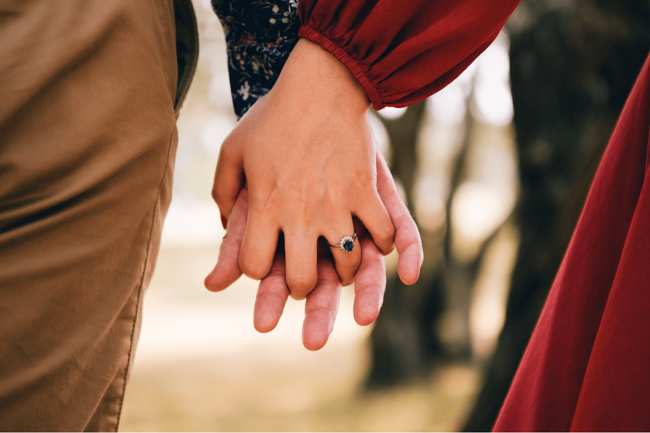 A couple holding hands, the woman wearing a sapphire, halo, oval cut engagement ring