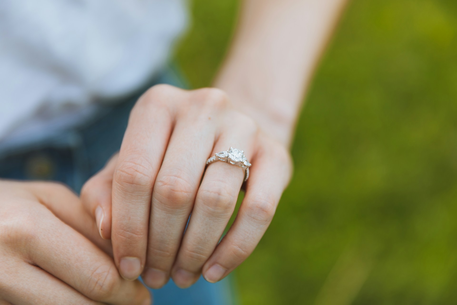 A woman demonstrates her engagement ring, which has a central diamond and two smaller side stones, against a green outdoor background.