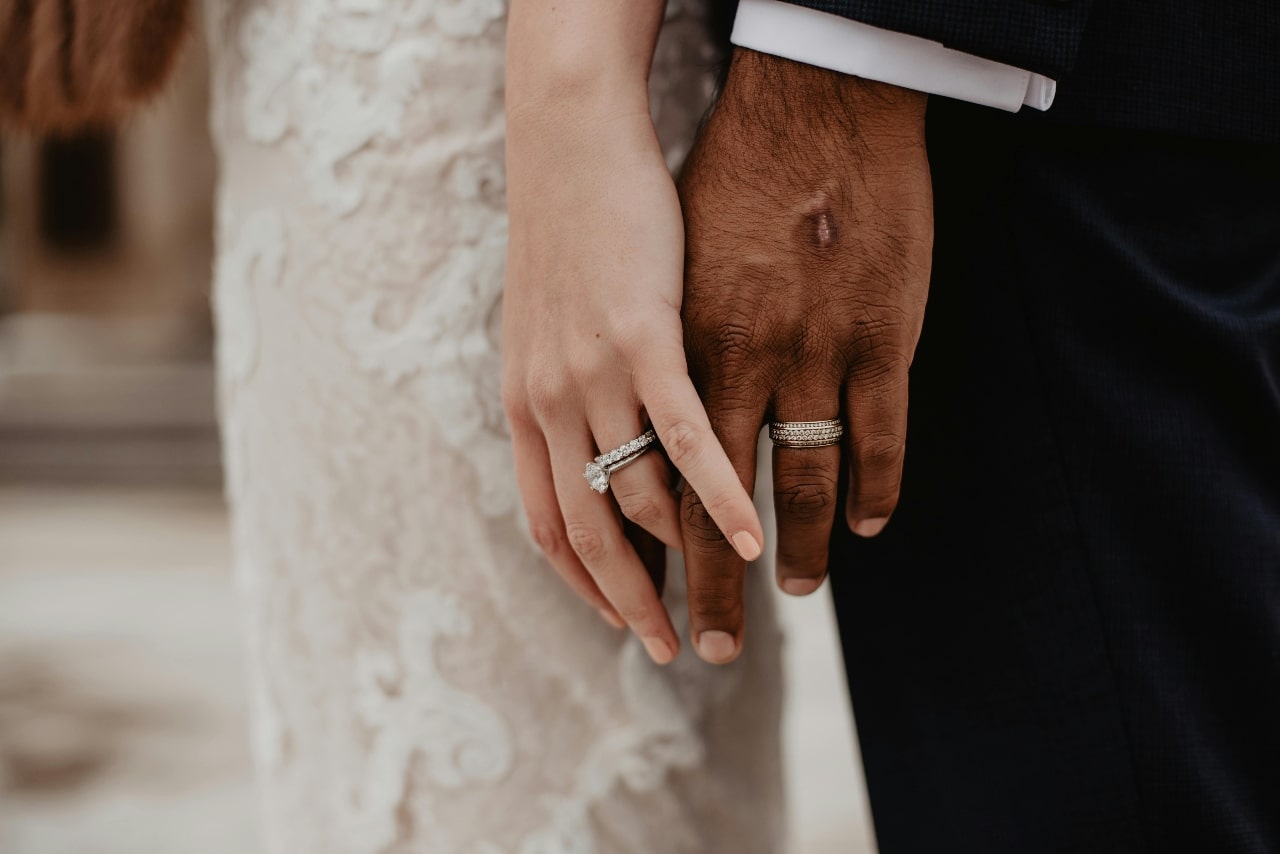 A close-up of a bride and groom holding hands, both wearing matching diamond wedding bands.