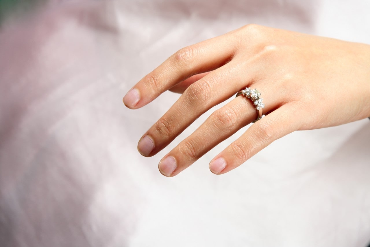 A close-up of a woman’s hand wearing a side stone engagement ring adorned with shining diamonds against a soft white background.
