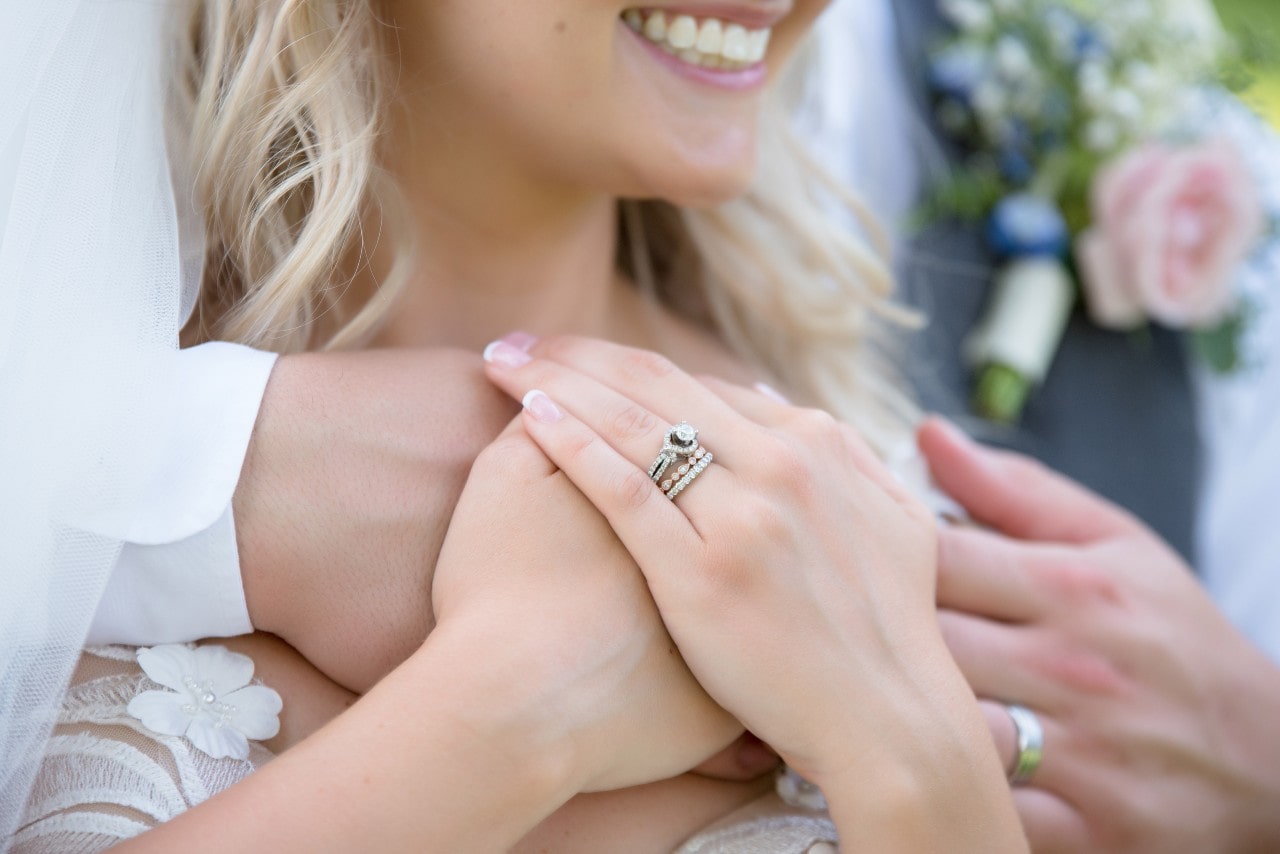 Close-up of a smiling bride holding hands with her partner, showcasing wedding rings with a bouquet with pink and blue flowers blurred in the background.