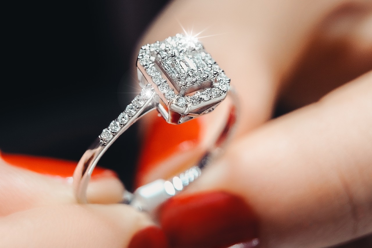 A close-up of a sparkling diamond ring held between fingers with red-painted nails.