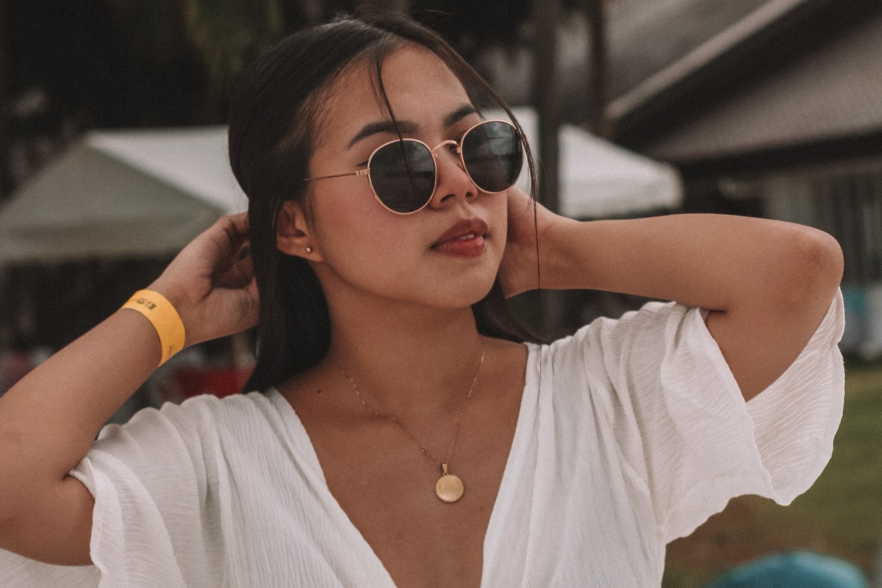 A woman at a festival wears a white tee, sunglasses, and a gold coin pendant.
