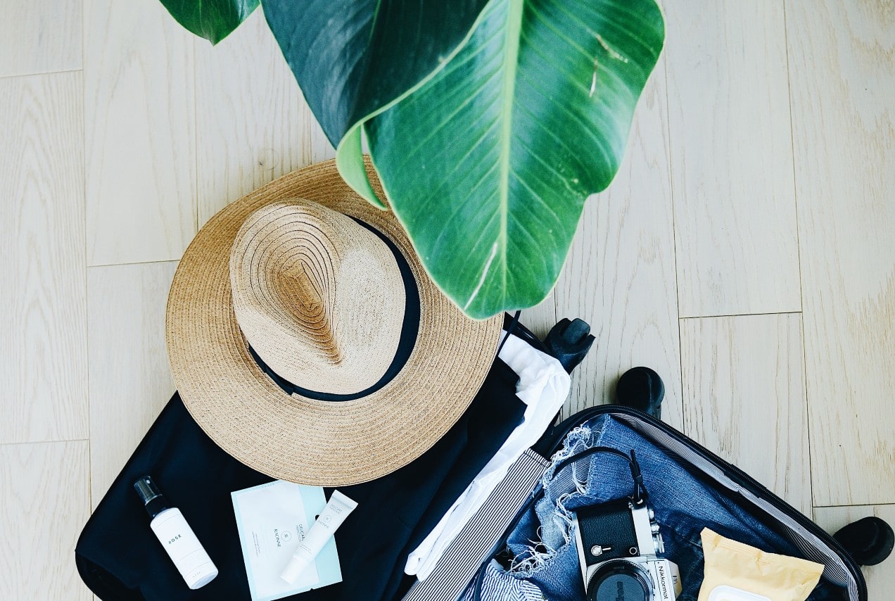 A suitcase packed with summer clothes and accessories sits under a house plant