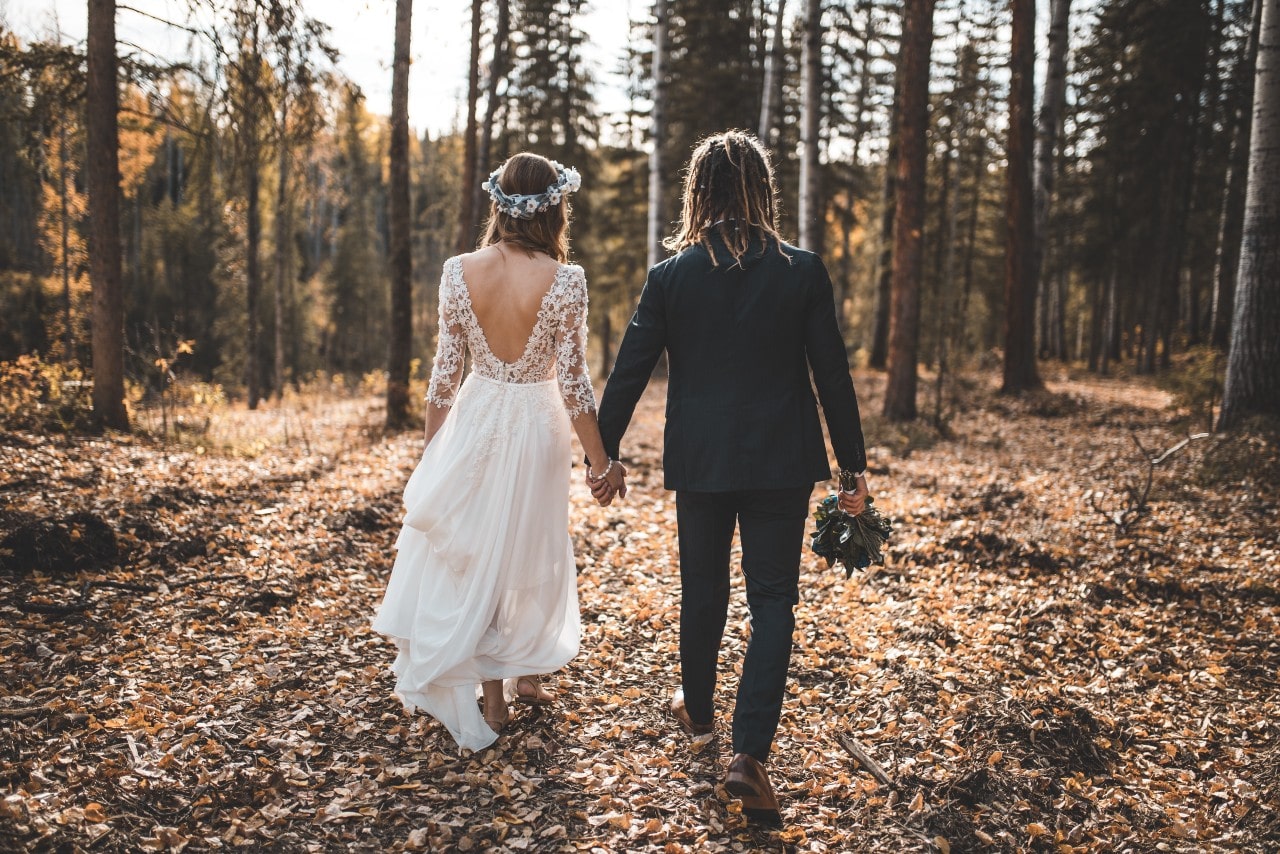 a bride and grooms holding hands and walking away from the camera in a forest during the fall