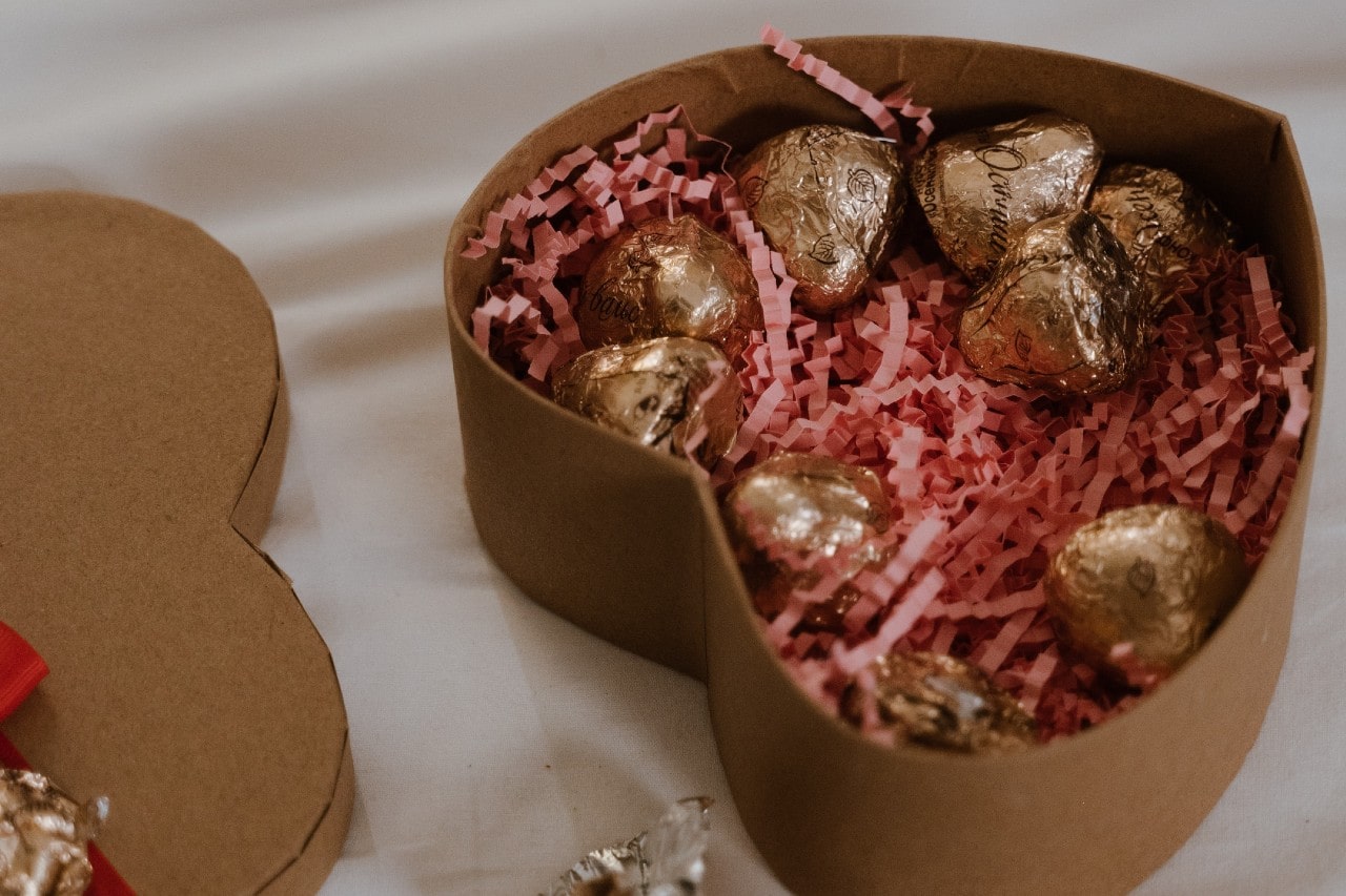 An array of chocolates in a heart-shaped cardboard gift box on beige sheets.