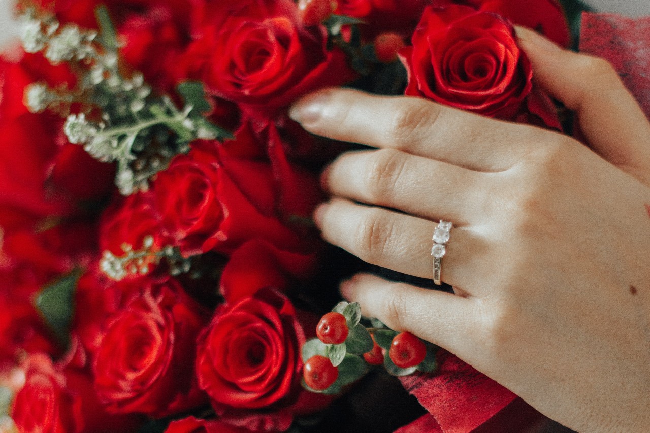 A close up of a three stone diamond engagement ring on a woman’s hand that is on top of a bouquet of red roses