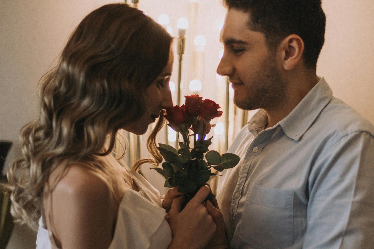 A couple celebrating Valentine’s Day together; the woman is smelling the red roses her boyfriend is holding