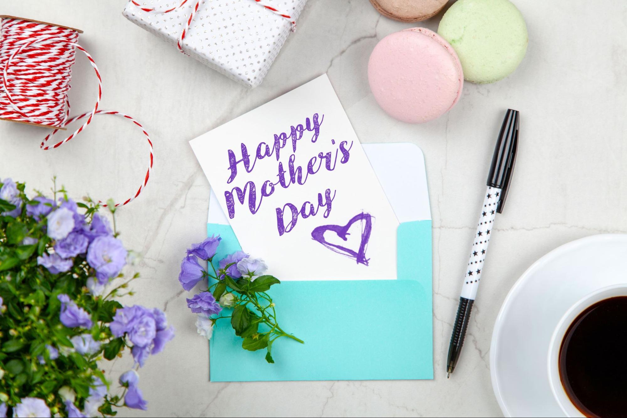 A Mother's Day card with "Happy Mother's Day" written in purple, placed in a blue envelope, surrounded by flowers, a pen, coffee, macarons, and a wrapped gift on a marble surface.