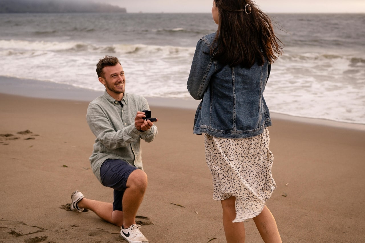 A man down on one knee proposing to a woman on the beach.
