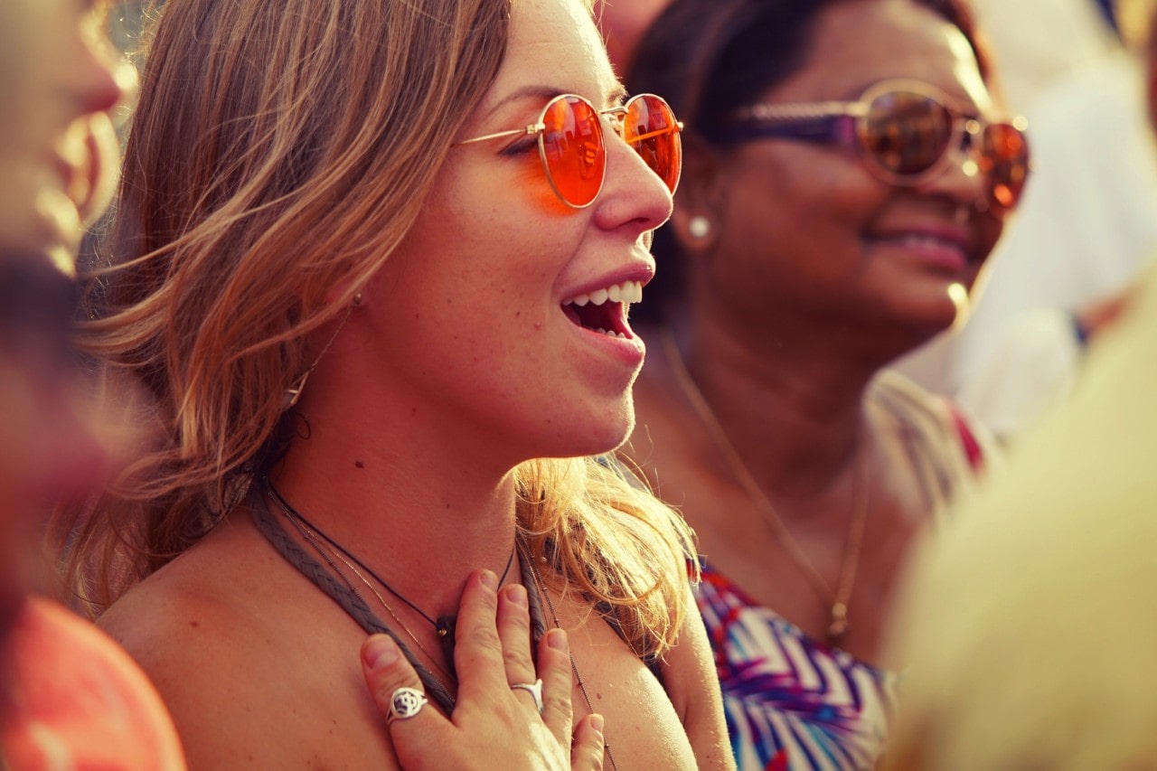 A woman wearing orange-tinted sunglasses smiles with her hand on her chest at an outdoor event, surrounded by a crowd.