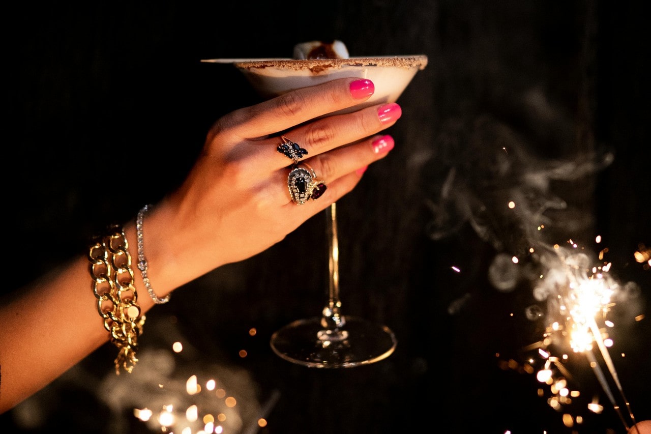 A woman's hand adorned with gold chain bracelets and gemstone rings holds a cocktail glass in front of festive sparklers.