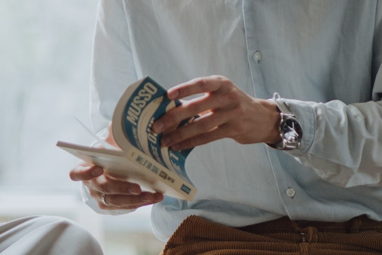 A person in a light blue shirt reads a book titled "Musso." They wear a silver watch, conveying calm focus. The background is softly blurred.