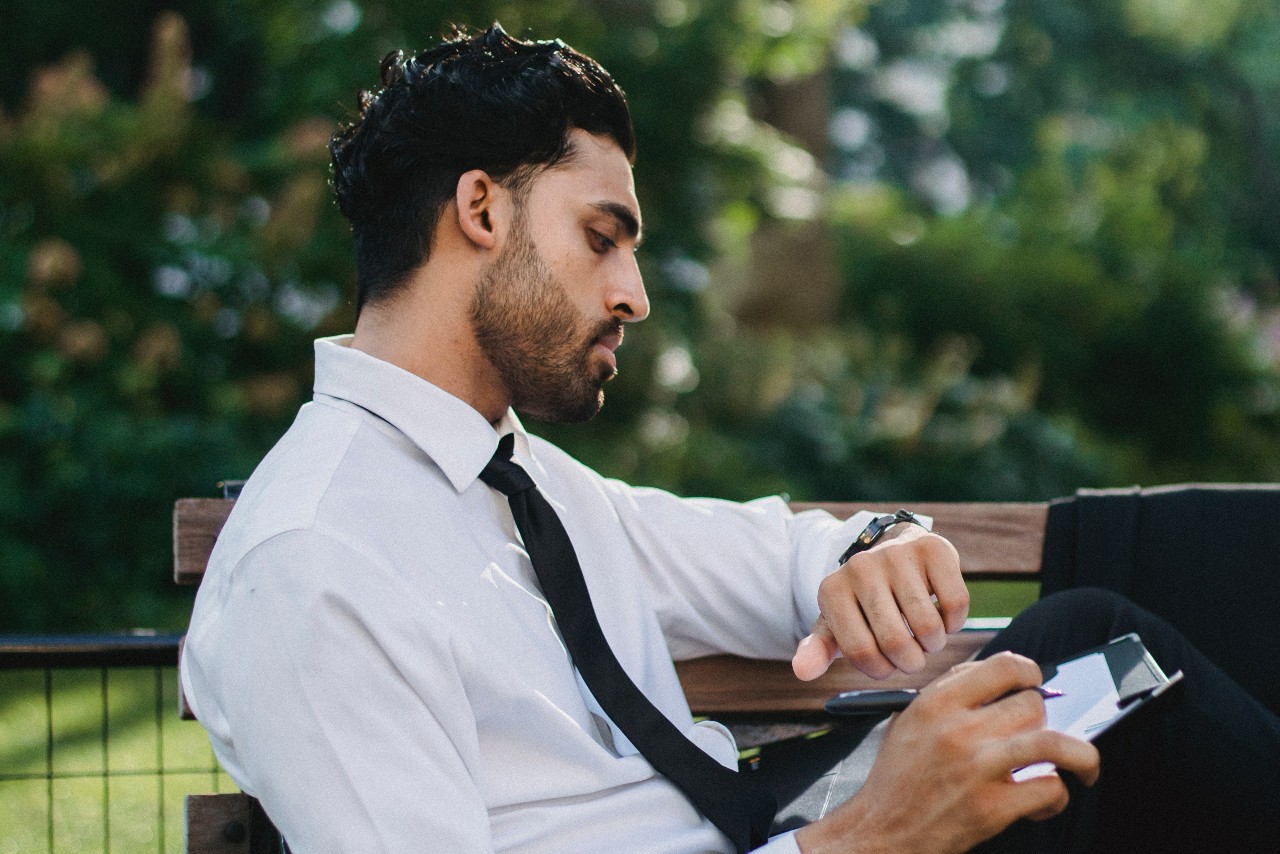 A man in a white shirt and black tie sits on a bench, checking his watch with a contemplative expression. He holds a pen and a notebook. Lush greenery in the background.