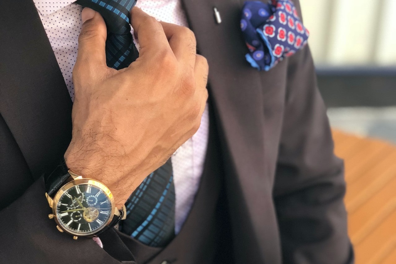 Close-up of a man in a brown suit adjusting a blue-striped tie. He wears a gold watch and a polka-dotted pocket square, conveying elegance.