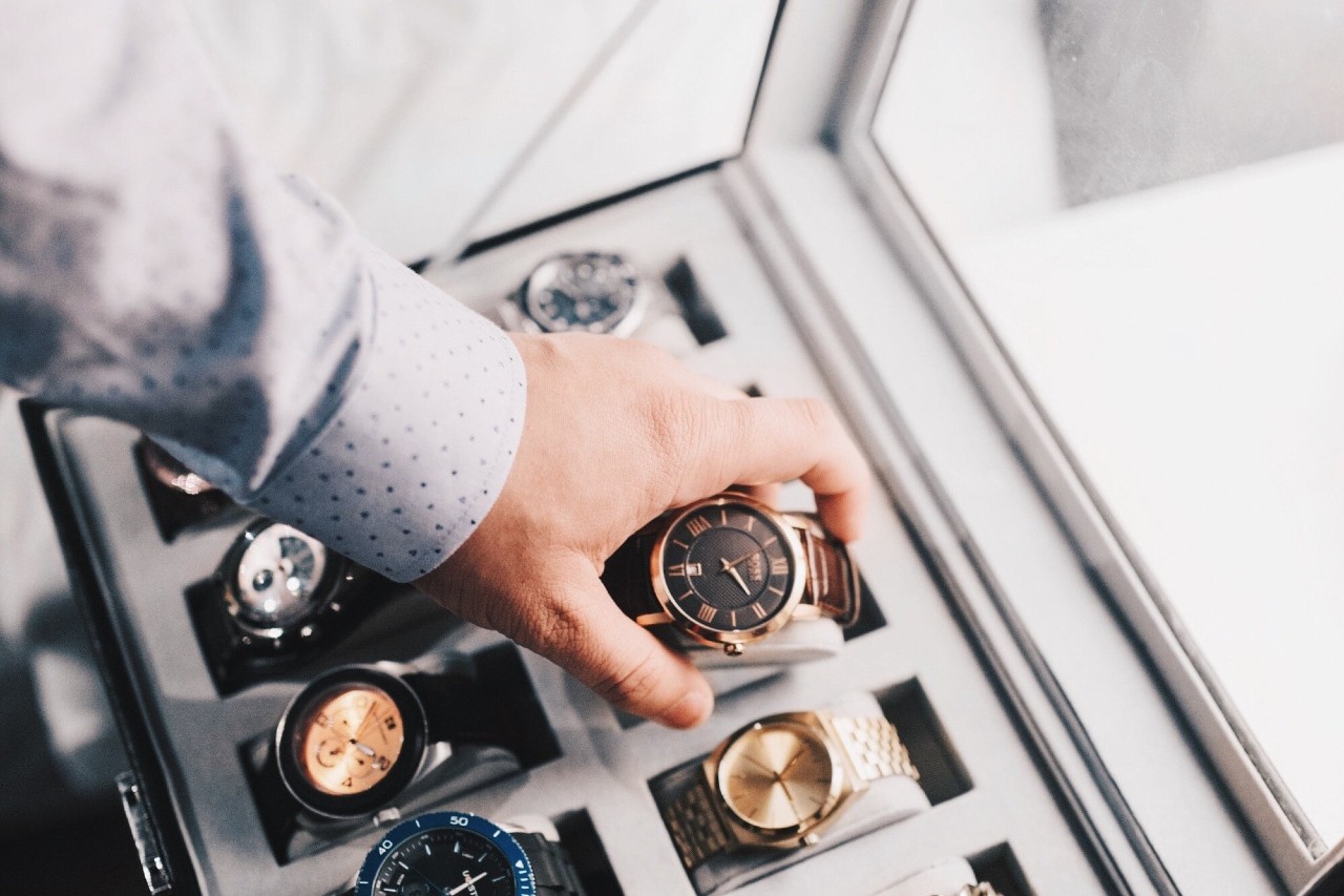 A hand in a polka dot shirt selects a luxury watch from a display case filled with different styles and colors, suggesting elegance and choice.