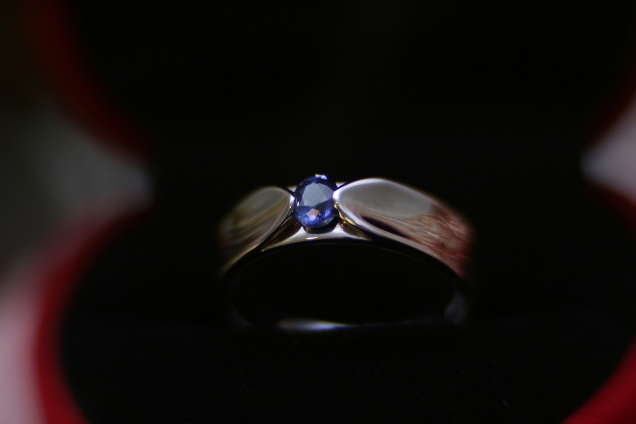 A platinum wedding band with oval blue sapphire as a centerpiece against a blurry background of an open jewelry box.