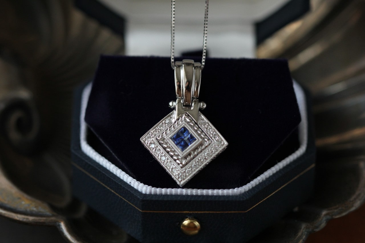 A white gold square shaped necklace with textured surface and four square sapphires at the center on a dark blue jewelry box.