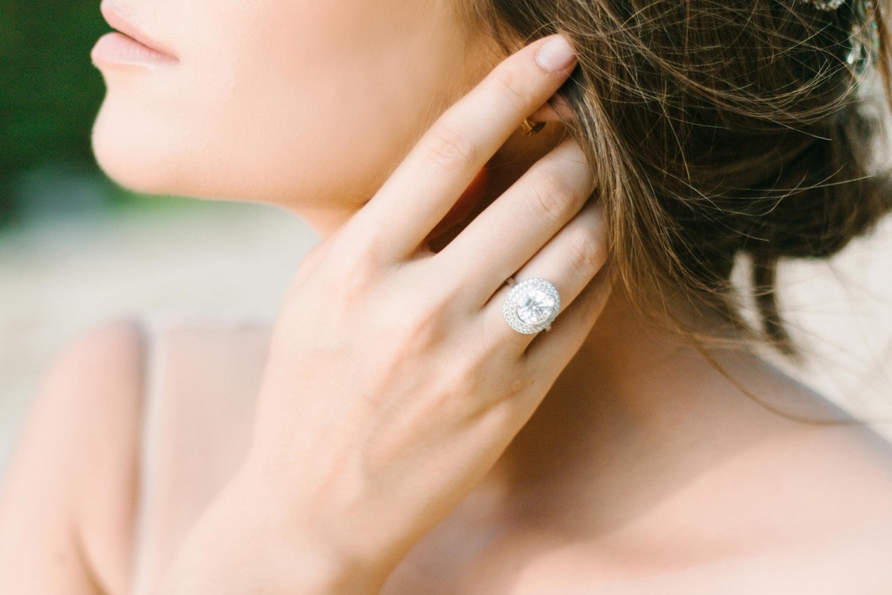 A close-up of a bride-to-be’s hand adorned with an ornate halo engagement ring, as she tucks a lock of hair behind her ear.