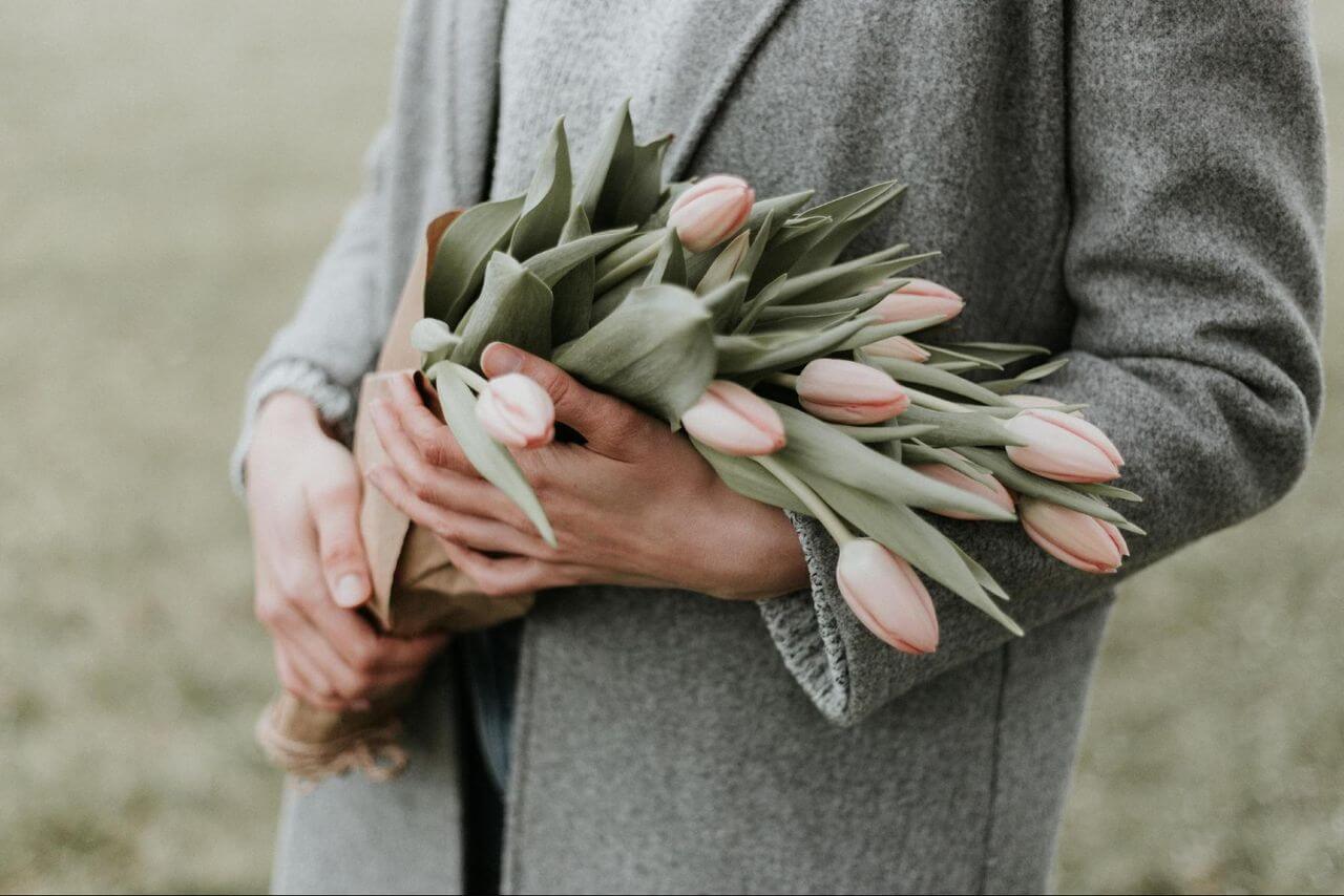 A man in a long grey coat holding baby pink tulips in an open field.