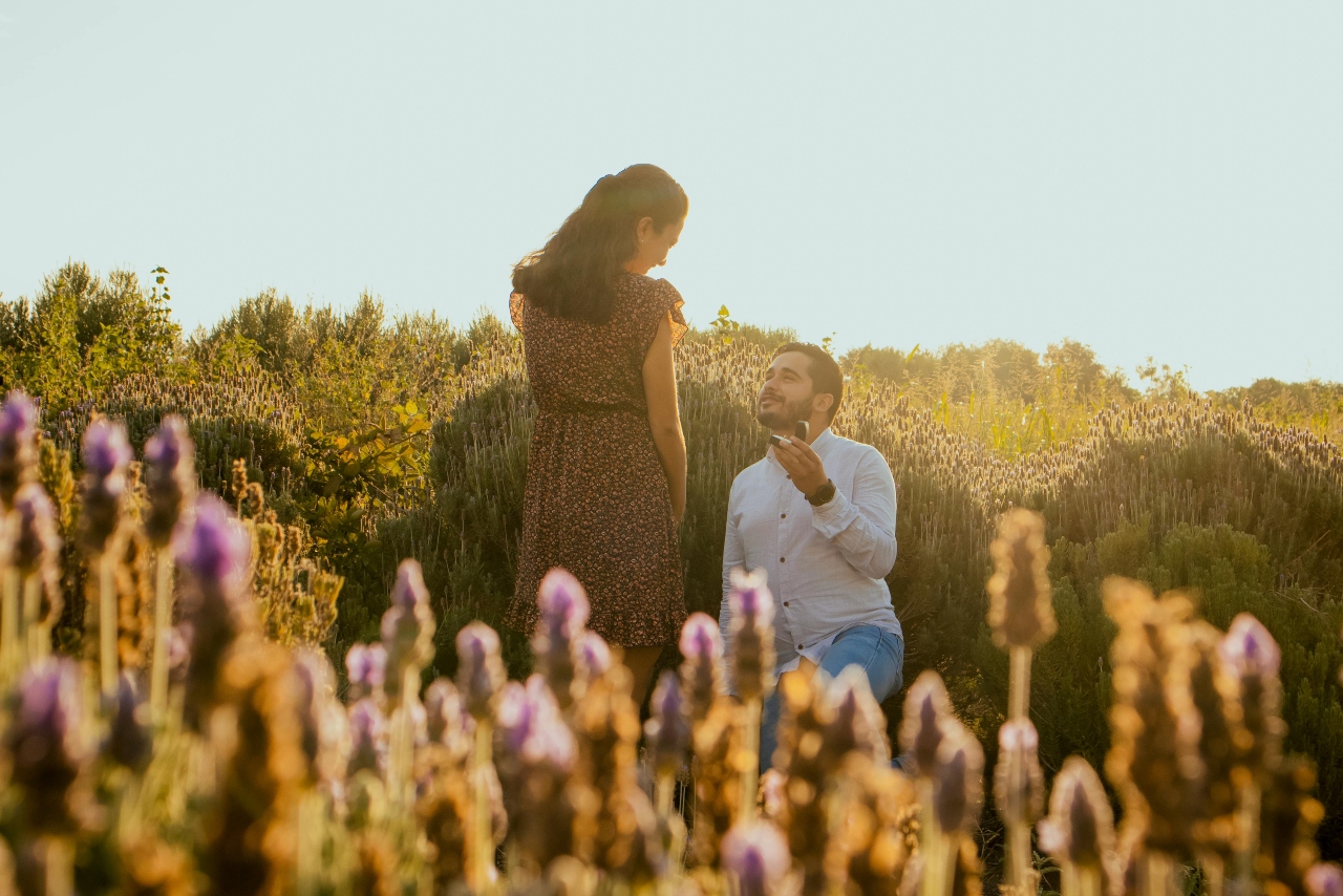 A couple in a beautiful garden with purple flowers, the man kneeling with an engagement ring.