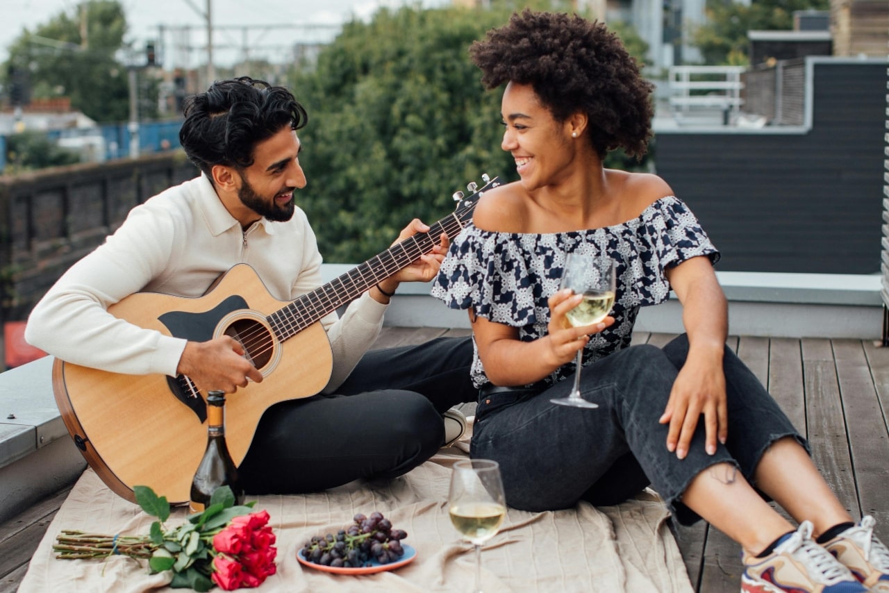 A couple enjoying a date on the rooftop with wine, roses and champagne, while a man is playing guitar for a woman.