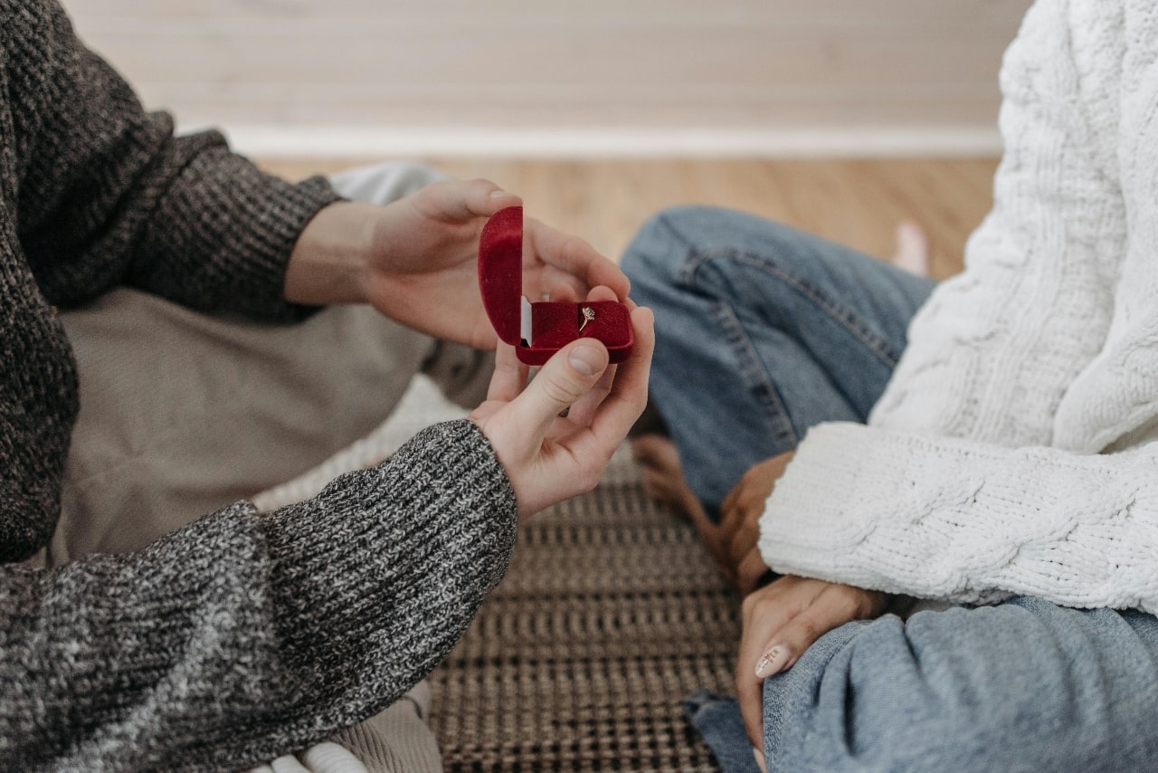 A heartfelt proposal moment captured indoors as one person presents an open red velvet ring box to their partner while sitting together in cozy sweaters and jeans.
