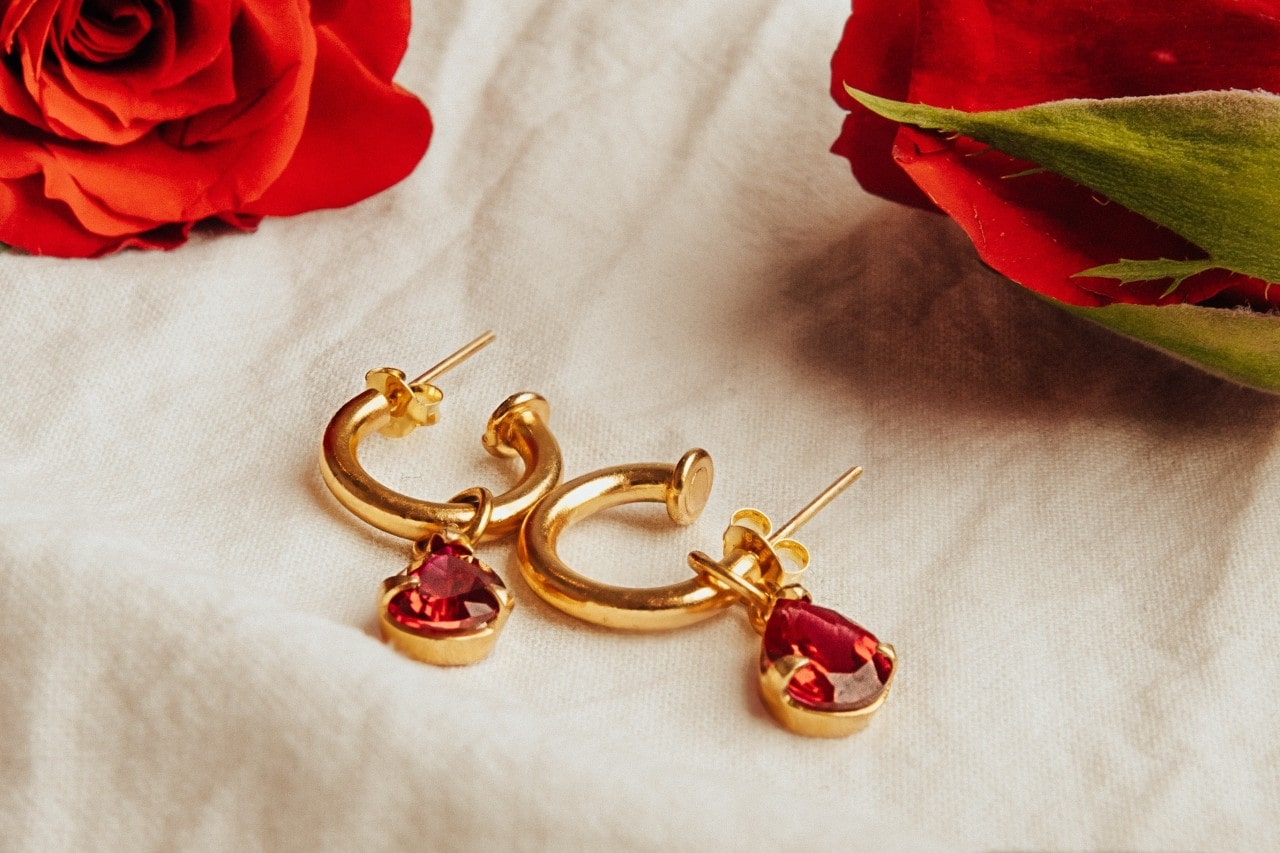 A pair of yellow gold ruby hoop earrings resting on a white cloth surrounded by red roses.