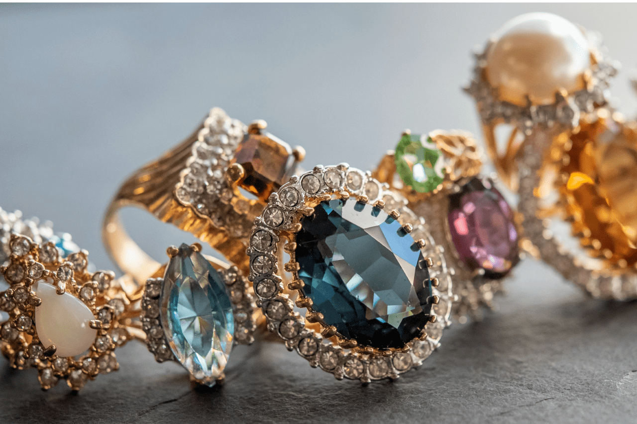 A collection of yellow gold gemstone fashion rings with diamond halos lay on black, faux leather fabric