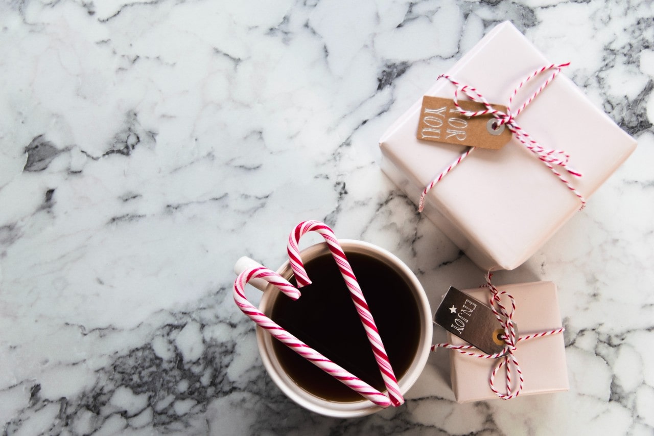 Two small, wrapped jewelry gifts accompany a mug of hot cocoa with two candy canes on a marble kitchen counter