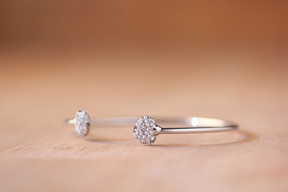 A close-up of a designer sterling silver cuff with diamond encrusted hearts on the ends sits on a cotton piece of peach-colored fabric